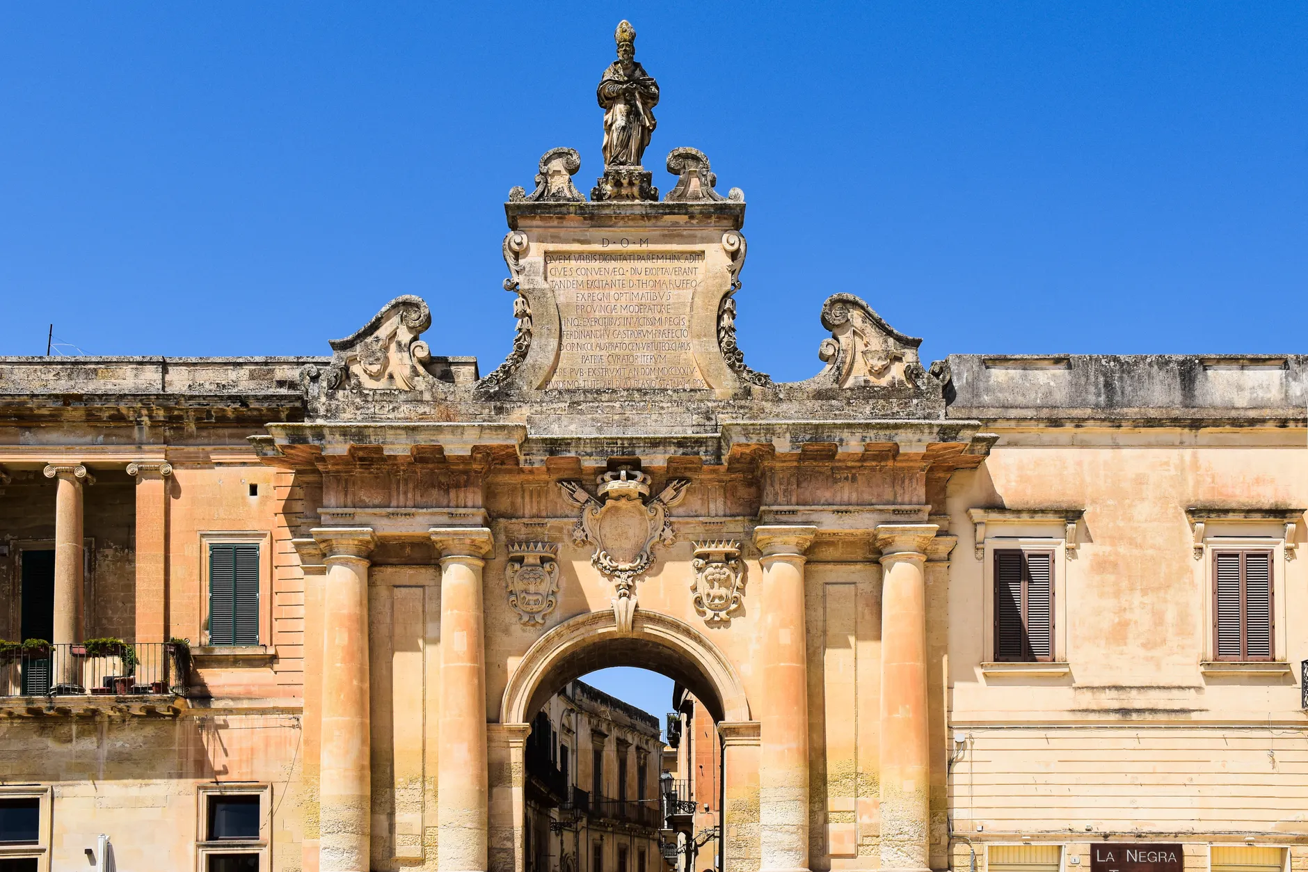 Ornate historical archway under a clear blue sky in Lecce, Apulia, Italy.