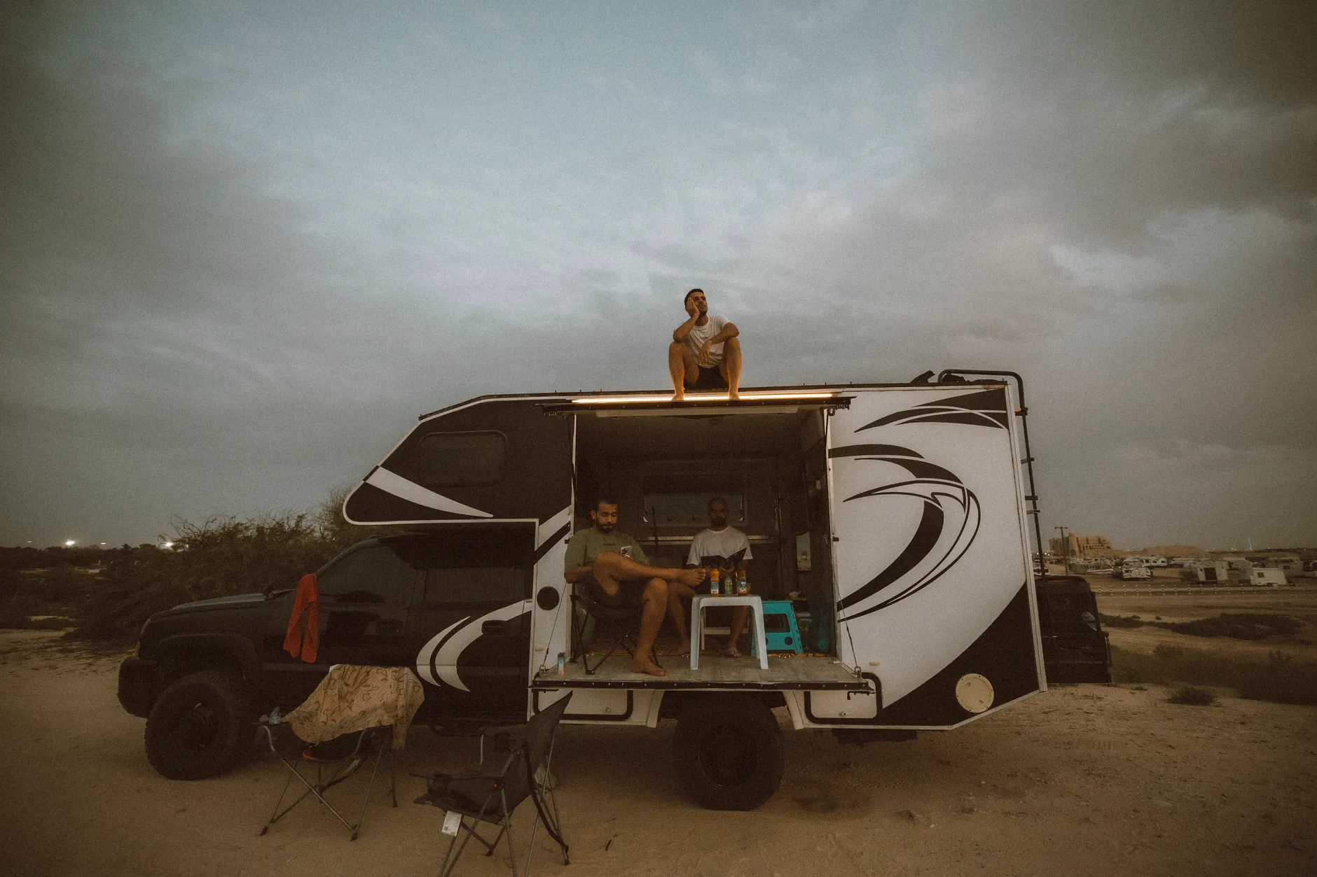 Three friends enjoying a camping trip in Dubai's desert, relaxing near a camper van.